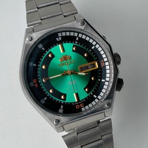 May include: A stainless steel Orient SK watch with a green and black dial. The watch features gold accents, a day-date display, and a metal band. The watch face displays the Orient logo and the text "SK Crystal 21 Jewels".