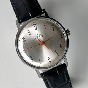 Könnte beinhalten: Eine silberfarbene analoge Armbanduhr mit rundem Zifferblatt, schwarzen arabischen Ziffern und einem dünnen roten Sekundenzeiger. Das Zifferblatt zeigt den kyrillischen Text "ЛУЧ" und "23 КАМНЯ" sowie "СДЕЛАНО В СССР" unten. Mit schwarzem Lederarmband.