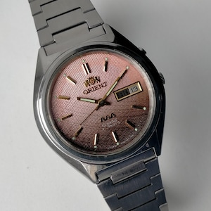 May include: A silver-toned Orient wristwatch with a salmon-colored dial. The watch features a day and date display, with the day reading "SAT 13". The watch has a silver metal band and a textured dial.