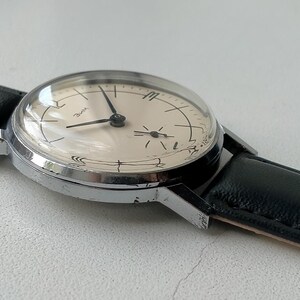 May include: A vintage wristwatch with a silver-colored case and a cream-colored face. The watch has black hands and a black leather band. The watch face has a domed crystal and is set against a light background.