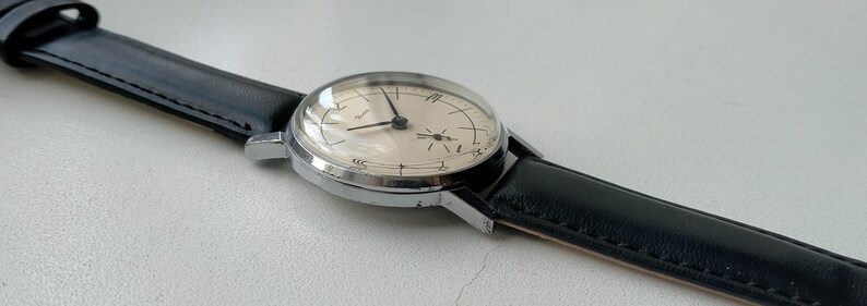 May include: A vintage wristwatch with a silver-colored case and a cream-colored face. The watch has black hands and a black leather band. The watch face has a domed crystal and is set against a light background.