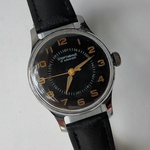 May include: A vintage wristwatch with a black face and gold-colored hands and numerals. The watch has a silver-colored case and a black leather band. The watch face includes the text "Спортивные 17 камней".
