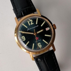 May include: A vintage wristwatch with a gold-tone case and a black leather band. The watch face is dark green with white numerals and hands. The text "Камандирские" and "чистополь" are visible on the watch face.