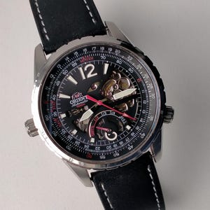 Könnte beinhalten: Schwarze Orient Automatik-Armbanduhr mit Skelett-Zifferblatt, silberfarbenen Zeigern und Stundenmarkierungen. Die Uhr hat ein schwarzes Lederarmband mit weißen Nähten und ein Edelstahlgehäuse. Das Zifferblatt enthält eine Tachymeterskala. Der Durchmesser beträgt ca. 40 mm.