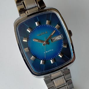 Puede incluir: Un reloj de pulsera plateado con una esfera cuadrada azul. La esfera del reloj tiene una ventana de fecha que dice "BC 4" y el texto "Чайка Кварц" en escritura cirílica.