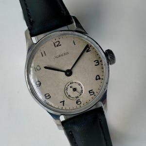 May include: A vintage wristwatch with a silver-tone circular case and a black leather strap. The off-white dial features black Arabic numerals, black hands, and a small sub-dial at the six o'clock position. The word ПОБЕДА is printed in black.