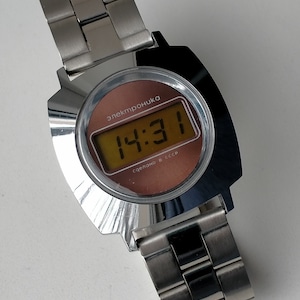 May include: A silver digital watch with a brown face and a silver metal strap. The watch face displays the time 14:31 and the text "электроника" and "сделано в ссср".