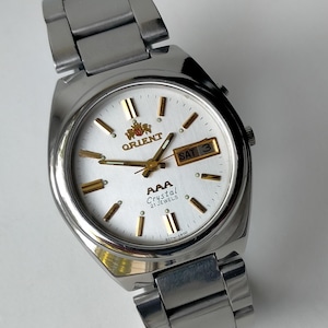 May include: A silver-toned stainless steel wristwatch with a white dial, gold-colored hour markers, and gold hands. The dial features the Orient logo, the text AAA Crystal 21 Jewels, and a day-date window displaying SAT 3 on the right side.
