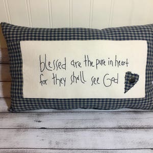 May include: Rectangular pillow with a blue and white gingham pattern. The center features a white rectangle with the handwritten text "blessed are the pure in heart for they shall see God." A heart-shaped patch and button are on the right.