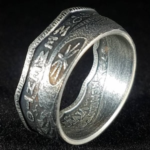 May include: A silver-toned ring with engraved text and floral designs. The ring has a wide band with a textured surface and a dark, oxidized finish. The inscription includes the words "Memento Vivere".