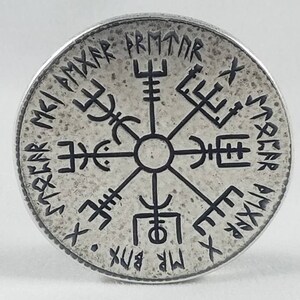 May include: A silver-colored circular pendant with a black compass design and runes around the edge. The runes read "MR. BIX. X.  SVEA.  R.  PMTAR.  X.  SMAR.  A.  VIX.  X.  MR.  BIX."