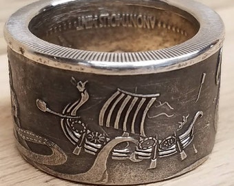 May include: A silver ring made from a coin featuring a Viking ship with a sail and oars, surrounded by a serpent. The coin's edge is visible around the ring.