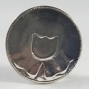 K&ouml;nnte beinhalten: Silberfarbener Metall-Token mit einem stilisierten Tulpenmotiv.