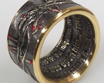 May include: A silver ring with a gold band, featuring a detailed design of a stylized tree with red accents. The ring is inscribed with the text "In God We Trust" and "Liberty 525"