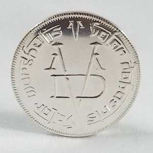 K&ouml;nnte beinhalten: Eine silberne M&uuml;nze mit einem erhabenen Design eines stilisierten Buchstabens "V" mit zwei nach oben und zwei nach unten zeigenden Pfeilen. Die M&uuml;nze tr&auml;gt den Text "V for Victory" am Rand.