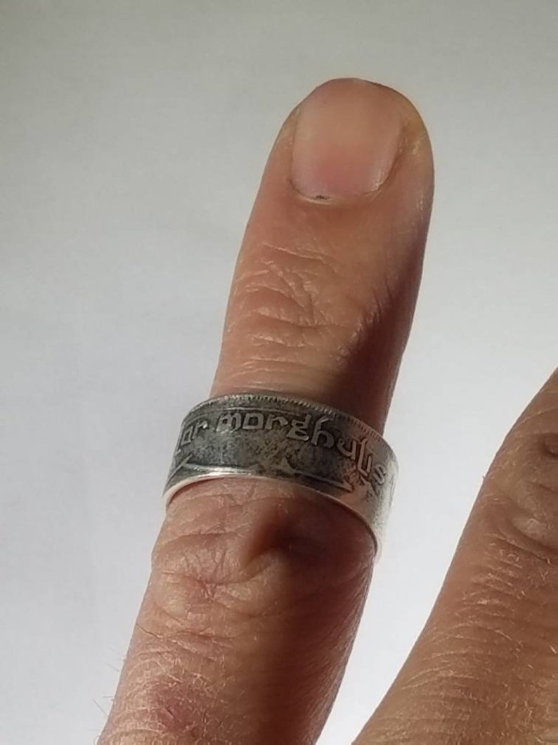 K&ouml;nnte beinhalten: Ein silberner Ring aus einer M&uuml;nze mit hebr&auml;ischer Schrift. Der Ring wird an einem Finger getragen.