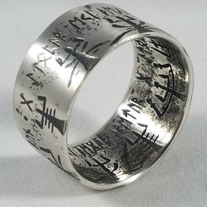 May include: A silver band ring with an inscription of runes around the entire band. The inscription is in a dark gray color.