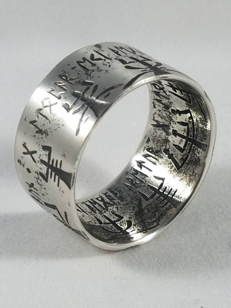 May include: A silver band ring with an inscription of runes around the entire band. The inscription is in a dark gray color.