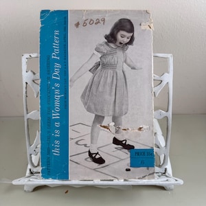 May include: Vintage sewing pattern for a girl's dress, featuring a black and white image of a young girl playing hopscotch. The pattern is titled "this is a Woman's Day Pattern" and includes the price of 35 cents.