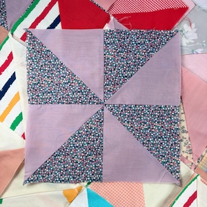 Op de afbeelding: Een vierkant quiltblok met een windmolenontwerp. De windmolen is gemaakt van lavendelkleurige stof en bloemenstof met kleine roze, witte en blauwe bloemen. De achtergrond toont andere quiltblokken met verschillende patronen en kleuren.