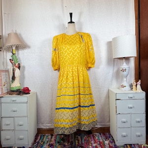 May include: A vintage yellow dress with short sleeves and a pleated skirt. The dress features a repeating pattern and is accented with blue stripes near the hem. The dress is displayed on a mannequin.