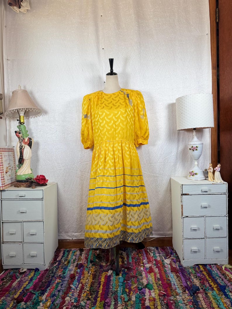 May include: A vintage yellow dress with short sleeves and a pleated skirt. The dress features a repeating pattern and is accented with blue stripes near the hem. The dress is displayed on a mannequin.