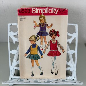 May include: Vintage Simplicity sewing pattern, size 3 child, featuring illustrations of three young girls in dresses. The pattern cover is red and white, with the Simplicity logo at the top. The pattern is displayed in a white metal decorative stand.