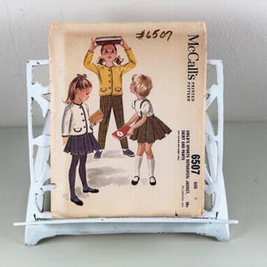 May include: Vintage McCall's pattern #6507 for children's sports separates, including a jacket, skirt, and pants. The cover features illustrations of three young girls in coordinated outfits. The pattern is displayed on a white metal stand.