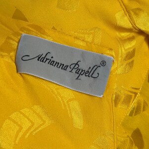 May include: Close-up of a yellow fabric with a subtle geometric pattern. A rectangular label with the text "Adrianna Papell" in elegant script is sewn onto the fabric. The label is light gray.