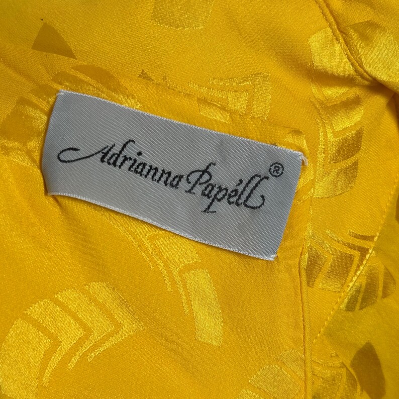 May include: Close-up of a yellow fabric with a subtle geometric pattern. A rectangular label with the text "Adrianna Papell" in elegant script is sewn onto the fabric. The label is light gray.