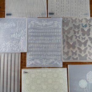 May include: A collection of eight rectangular plastic embossing folders featuring various textures and patterns, including musical notes with the text 'O COME ALL YE FAITHFUL', butterflies, poinsettias, stripes, and geometric floral designs.