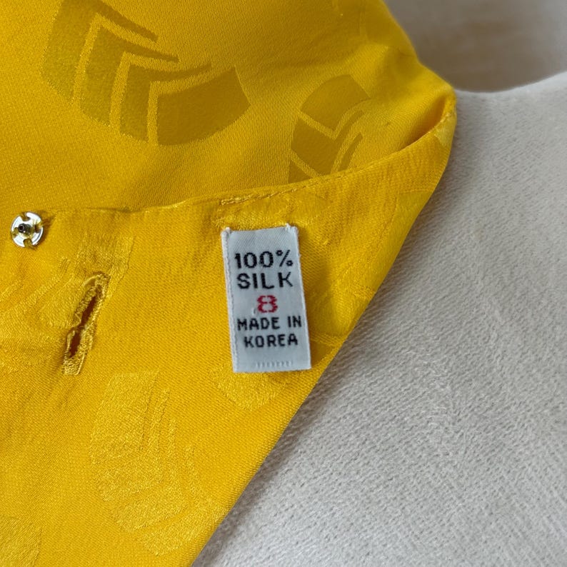 May include: Close-up of a bright yellow silk garment with a subtle geometric pattern. The label reads "100% SILK" and "MADE IN KOREA". The garment has a button and buttonhole detail.