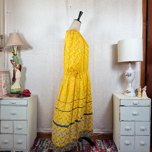 May include: A vintage yellow dress with a leaf pattern and blue stripes. The dress is displayed on a mannequin. The dress has long sleeves and a gathered waist. The hemline features a patterned border. The dress is a bright, cheerful color.