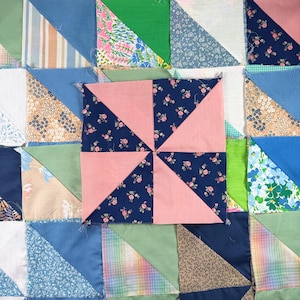 Op de afbeelding: Close-up van een patchworkquilt met een windmolenontwerp. De quilt heeft vierkanten in verschillende kleuren en patronen, waaronder bloemenprints, strepen en effen kleuren. De centrale windmolen is gemaakt van roze en marineblauwe stof.