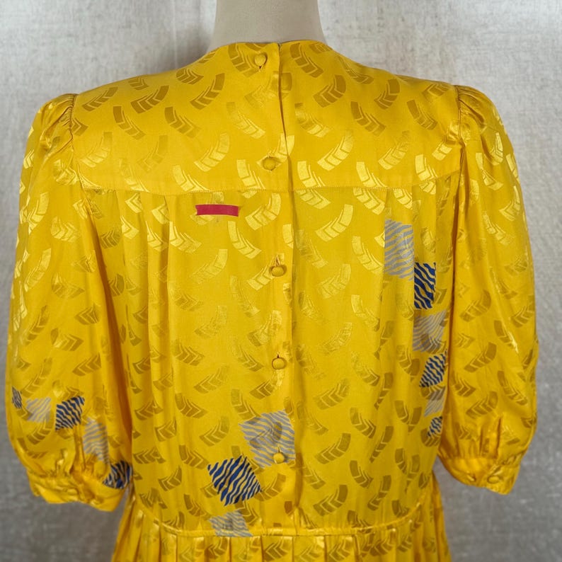 May include: A vintage yellow dress with a repeating pattern of abstract shapes. The dress features a button-down back, short sleeves, and a red horizontal stripe detail. Accents of blue and gray squares are visible.