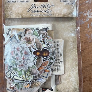 May include: A clear plastic package containing 134 pieces of paper ephemera labeled Tim Holtz idea-ology Field Notes. The collection includes various die-cut images of a large bumblebee, floral arrangements, butterflies, and insects on aged paper.