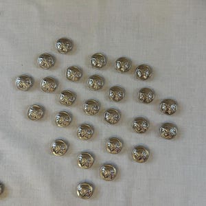 May include: A collection of silver-toned buttons with a raised design. The buttons are arranged in a scattered pattern on a white surface.