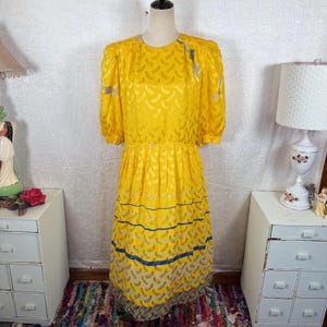 May include: A vintage yellow dress with a textured pattern and short, puffed sleeves. The dress features a fitted waist and a skirt with horizontal stripes in blue, gray, and a patterned design. The dress is displayed on a mannequin.