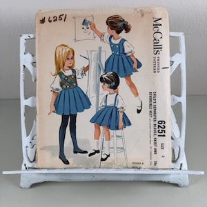 May include: Vintage McCall's pattern #6251 for child's separates, including a blouse, skirt, and reversible vest. The illustration shows three young girls in blue dresses. The pattern is displayed on a white metal stand.