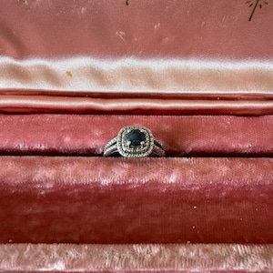 May include: A silver ring with a dark blue oval-shaped gemstone surrounded by smaller diamonds. The ring is displayed in a pink velvet ring box.
