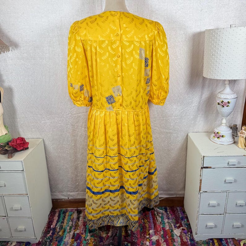 May include: A vintage yellow dress with a textured pattern and short, puffed sleeves. The dress features a button-up back, a fitted waist, and a patterned skirt with blue and gray accents. The dress is knee-length.