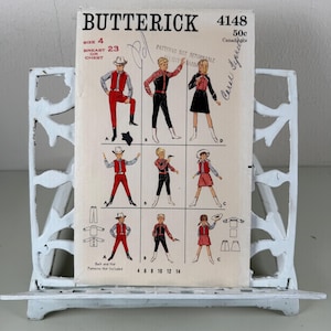 May include: A vintage Butterick sewing pattern, size 4, featuring illustrations of children's cowboy-themed outfits. The pattern includes a variety of designs with red, black, and white color schemes. The pattern is held in a white metal bookend.