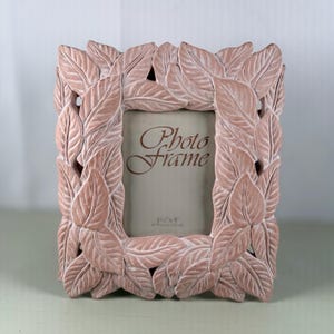 May include: A decorative photo frame with a leaf design in a soft, muted pink color. The frame is rectangular with a central opening for a 3.5 x 5 inch photo. The words "Photo Frame" are printed on the photo area.