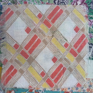 May include: A square fabric patch with a geometric pattern in red, yellow, and tan on a white background. The design features diagonal lines and rectangular shapes. The edges are frayed, and the patch is part of a larger textile.