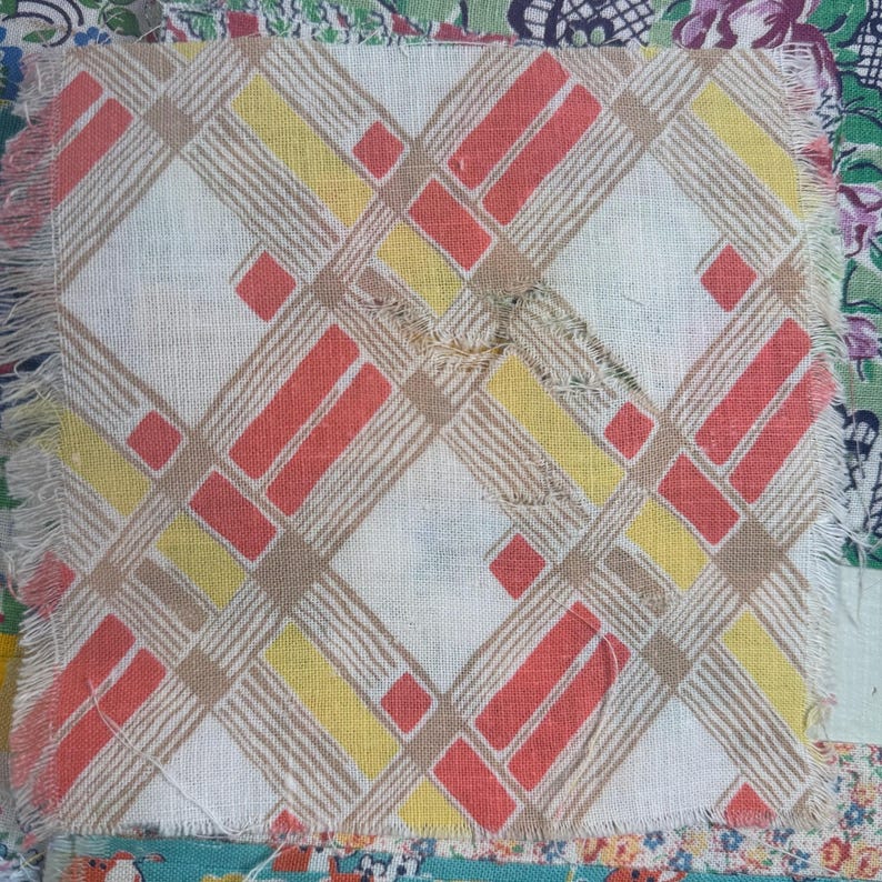 May include: A square fabric patch with a geometric pattern in red, yellow, and tan on a white background. The design features diagonal lines and rectangular shapes. The edges are frayed, and the patch is part of a larger textile.
