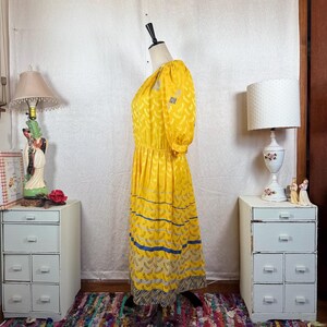 May include: A vintage yellow dress with a floral pattern and blue stripes. The dress has short sleeves and a gathered waist. The hemline features a patterned border. The dress is displayed on a mannequin.