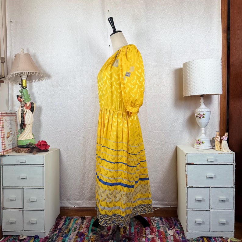 May include: A vintage yellow dress with a floral pattern and blue stripes. The dress has short sleeves and a gathered waist. The hemline features a patterned border. The dress is displayed on a mannequin.