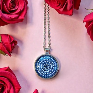 May include: A silver-toned necklace with a round pendant featuring a blue and white heart pattern. The necklace is displayed on a pink background, surrounded by vibrant red roses.
