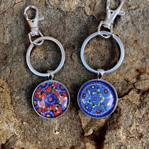 May include: Two silver keychains with round, colorful floral designs. The keychains have a silver clasp and a silver ring.