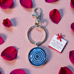 May include: Silver-tone keychain with a round, blue pendant featuring a white heart pattern. The keychain includes a clasp and a keyring. A small card with red trim and text is tied with a red ribbon. The background is pink with scattered red rose petals.